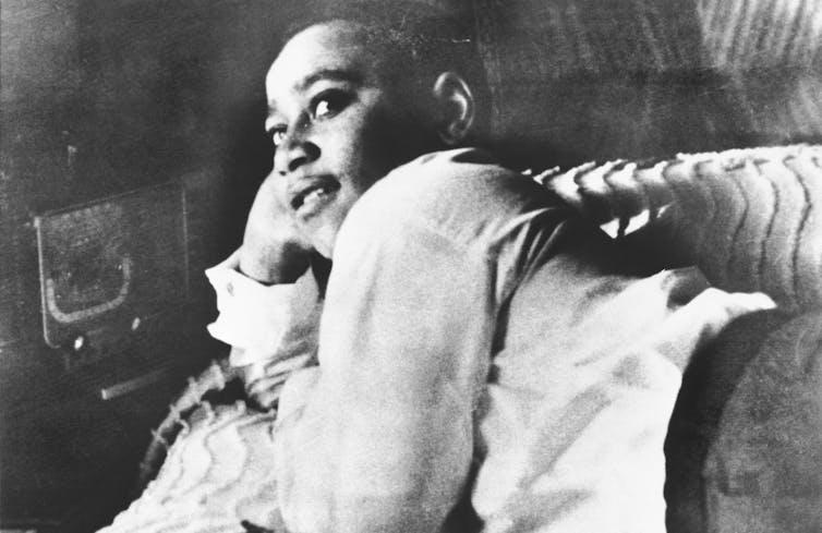 A young Black boy leans against his arm and reclines on a bed in a black and white photo.