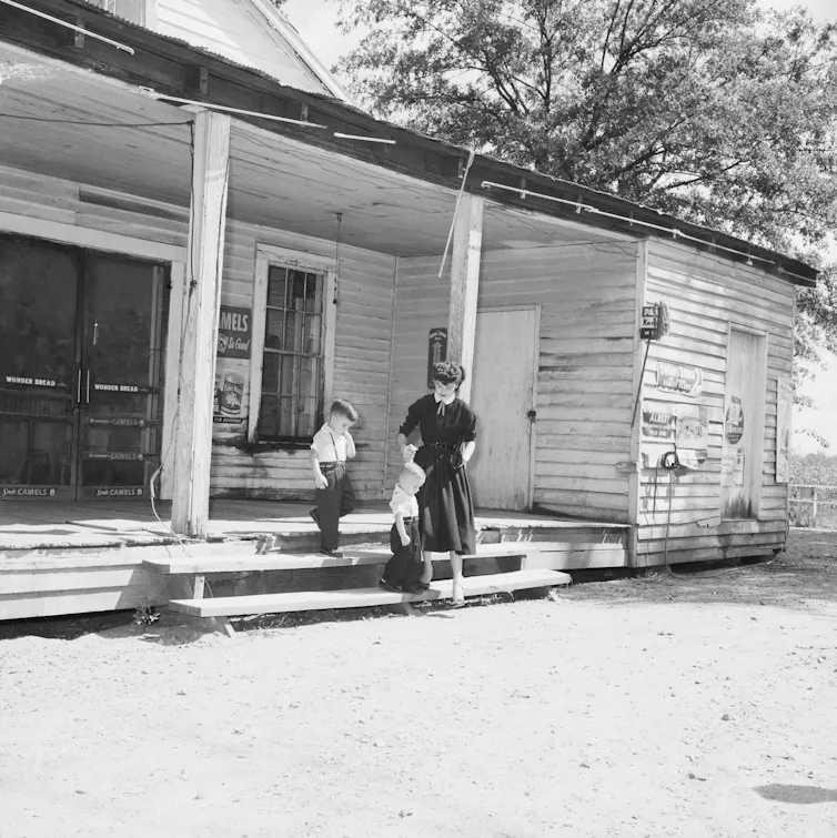 A woman stands with two young boys on the steps of a dilapidated looking wooden building.