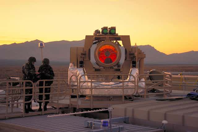 A photo of two soldiers standing next to a prototype laser weapons with a large orange lens.