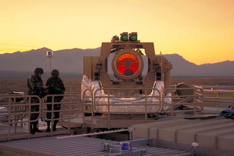 A photo of two soldiers standing next to a prototype laser weapons with a large orange lens.