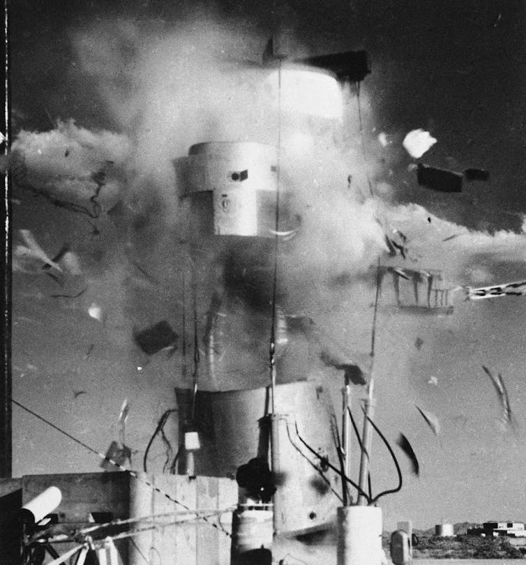 A black and white photo of a metal missile shell on a stand, appearing to explode.