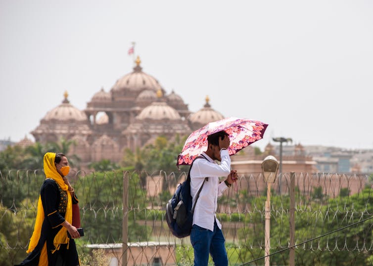 Two people, one with an umbrella, walking on a hot day with a Hindu temple in the background.