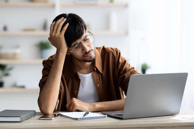 A stressed man wearing glasses sits at a desk looks at a laptop screen.