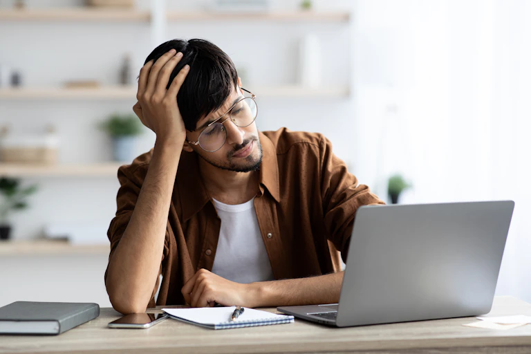 A stressed man wearing glasses sits at a desk looks at a laptop screen.