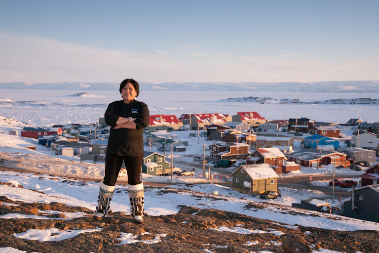 A woman wearing black standing on a hill. A snowy village is seen in the background.