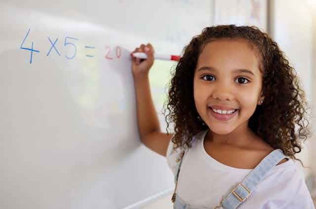 A girl faces the viewer as she writes a math problem on a whiteboard.