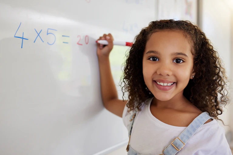 A girl faces the viewer as she writes a math problem on a whiteboard.