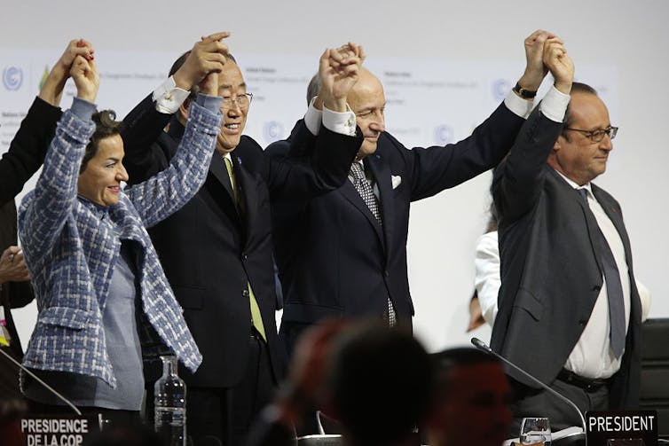 Three men in suits and one woman in a blue blazer hold raised hands triumphantly.