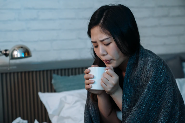 Young woman with a blanket wrapped around her holds a warm drink in her hands
