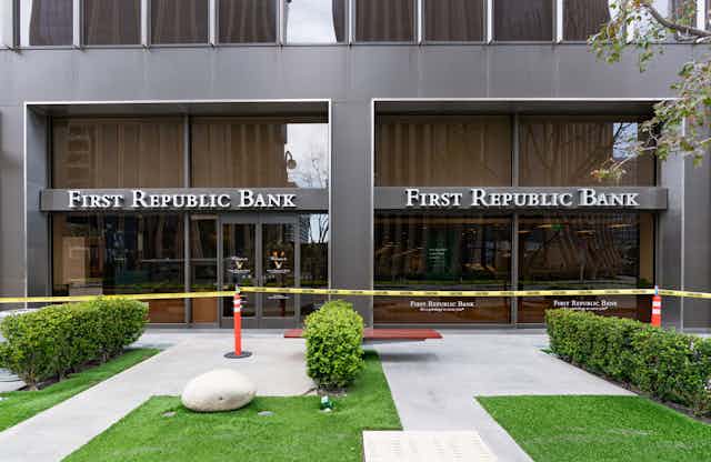 A gray building with the words first republic bank sits in front of a grass, bushes and sidewalk while yellow caution tape stretches across the building