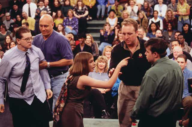 Woman points and yells at man while security intervenes on stage during taping of TV talk show.