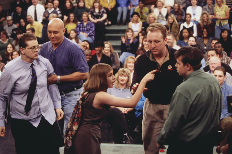 Woman points and yells at man while security intervenes on stage during taping of TV talk show.