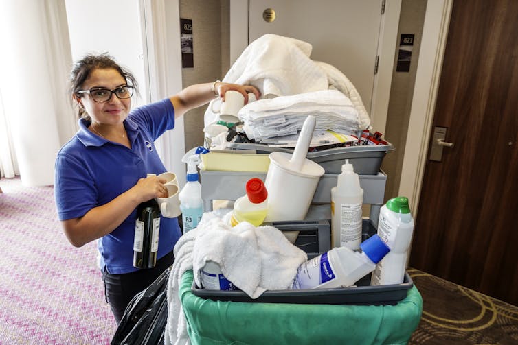 A hotel housekeeper stands next to her cart, piled with towels and bottled cleaning supplies.