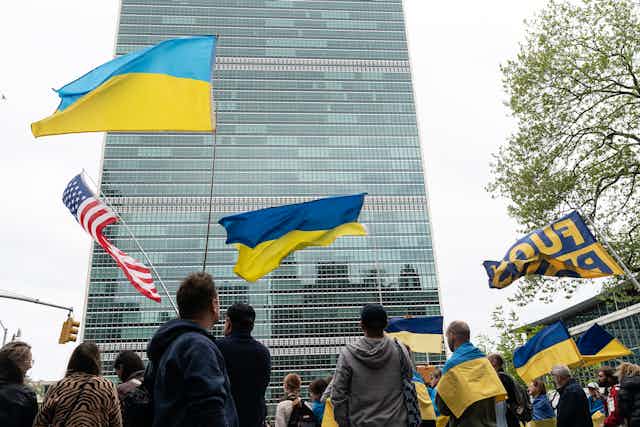 The backs of people are seen holding Ukrainian and American flags in front of a tall glass building. 