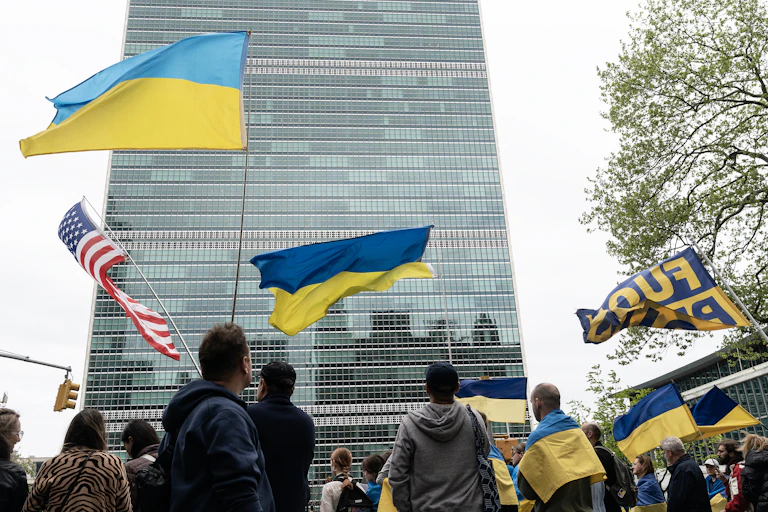 The backs of people are seen holding Ukrainian and American flags in front of a tall glass building.