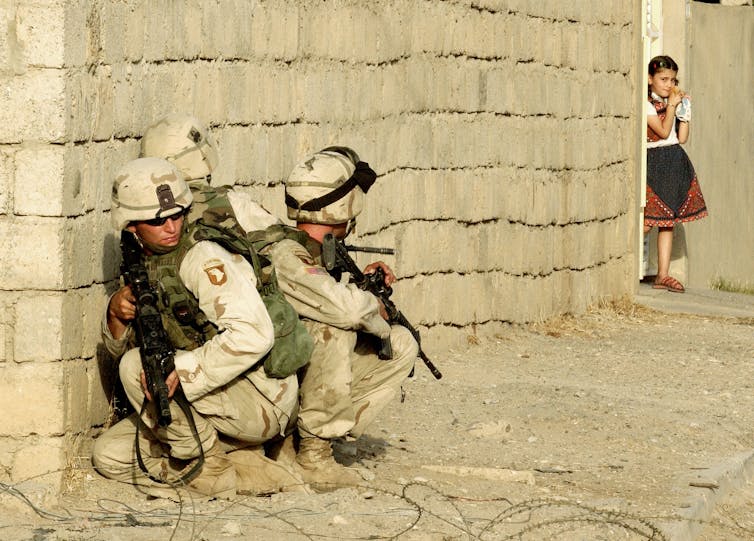 Soldiers in beige uniforms crouch down against a beige brick wall, while a young girl peers around the corner and watches them.