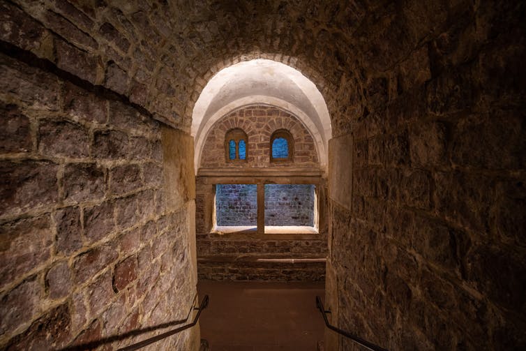 Stone steps in a high, vaulted, narrow passageway lead to arched windows with sunlight streaming in.