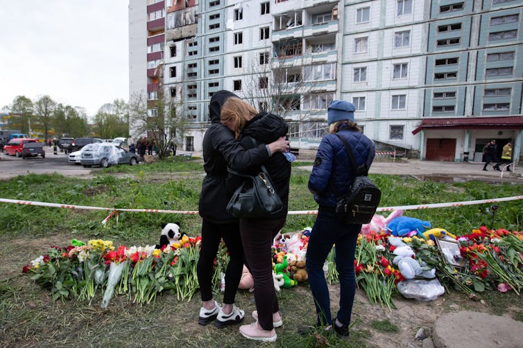 People embrace in front of flowers and teddy bears, in front of a building that looks partially charred.