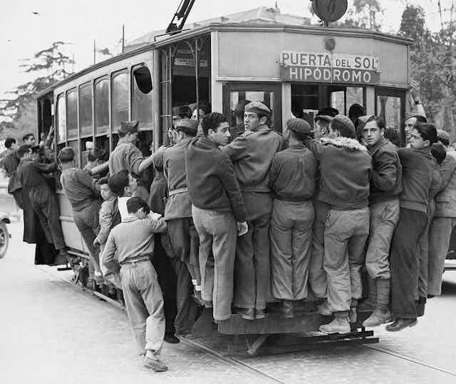 A black and white photo shows men in pants and informal jackets trying to ride on the outside of a trolley car.
