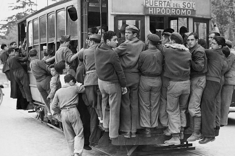 A black and white photo shows men in pants and informal jackets trying to ride on the outside of a trolley car.