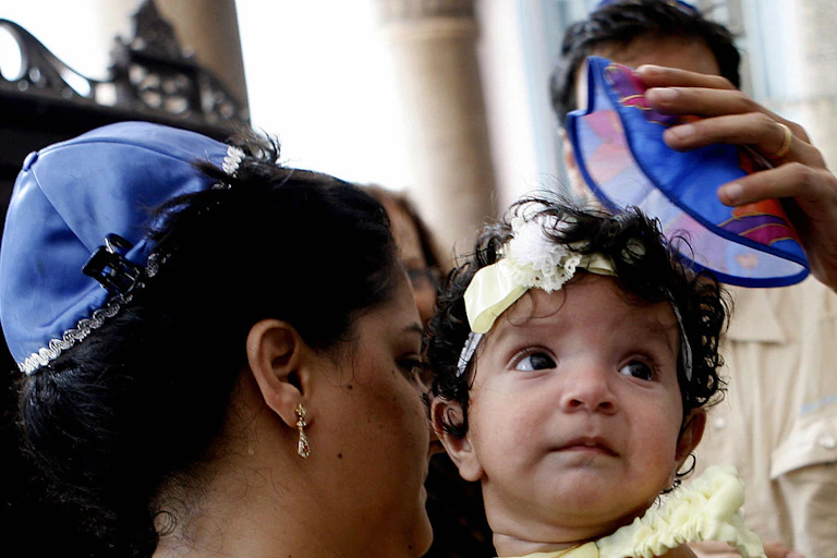 A woman wearing earrings and a blue skullcap holds a baby in a yellow outfit and yellow headband.
