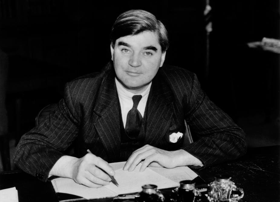A black and white photo of Aneurin Bevan who is at a desk writing with a pen in hand. He is staring at the camera. 