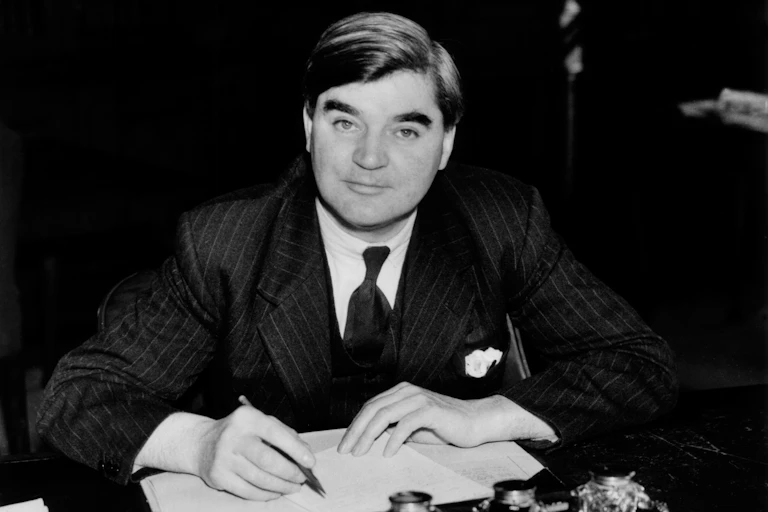 A black and white photo of Aneurin Bevan who is at a desk writing with a pen in hand. He is staring at the camera.