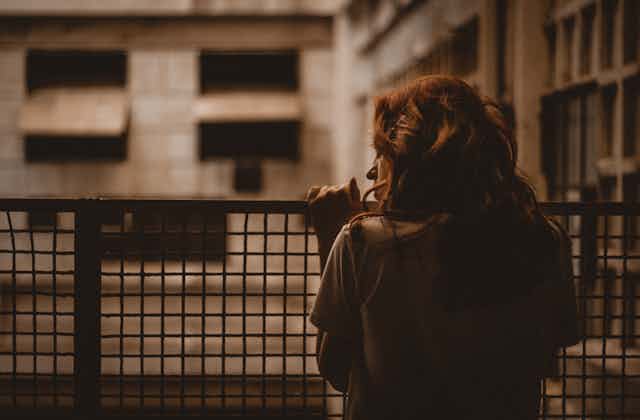 a woman with wavy red hair looks out at an apartment building from her wire-fenced balcony