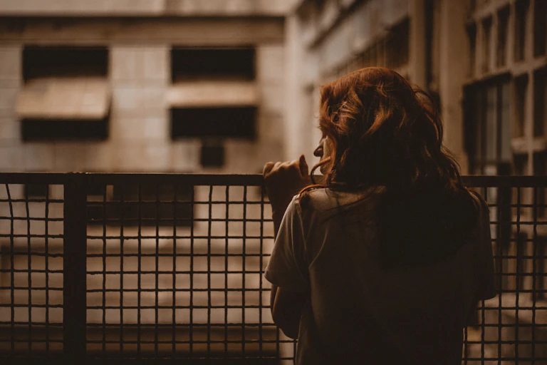 a woman with wavy red hair looks out at an apartment building from her wire-fenced balcony