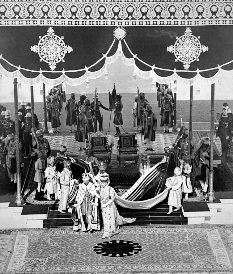 Black and white historic photo of the king and queen in full coronation regalia, surrounded by Indian princes and British Raj officials, under a ceremonial canopy, with ornate Indian-style mosaics on the ground