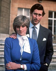 Diana usa uma jaqueta azul e Charles está atrás dela em um terno cinza escuro.