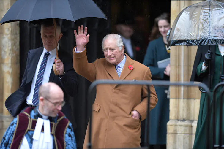 Charles waves from a step, an umbrella is held over him. He wears a camel coloured coat.