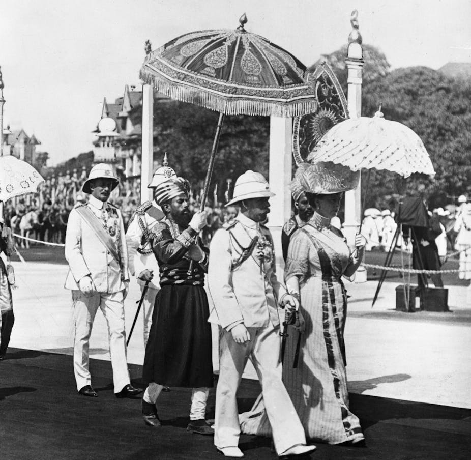 black and white historical photo of George V in a plinth helmet and Queen Mary walking in Bombay. An Indian man in uniform and turban is walking behind the royal couple, holding an ornamental parasol above the king's head