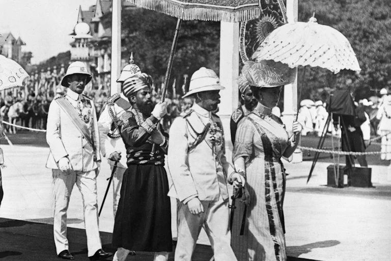 black and white historical photo of George V in a plinth helmet and Queen Mary walking in Bombay. An Indian man in uniform and turban is walking behind the royal couple, holding an ornamental parasol above the king's head