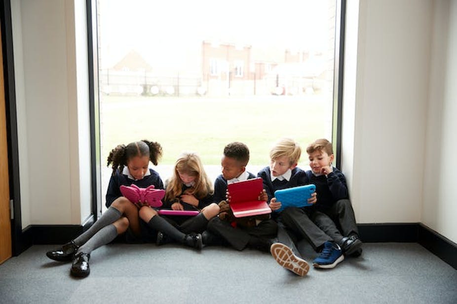 Cinq jeunes enfants en uniforme scolaire sont assis ensemble et regardent les écrans de leurs tablettes.