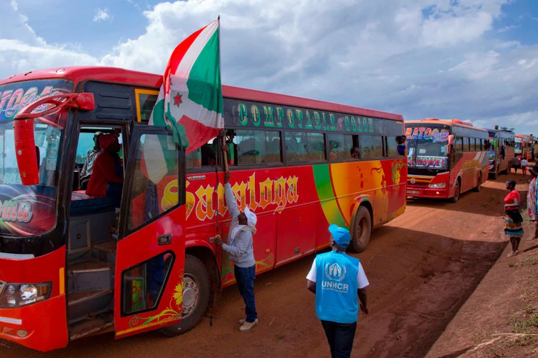 Man touching a wooden pole attached to a red, green and white flag that is strapped to a bus window. Three other stationary buses are behind it and a handful of people are standing along the road.