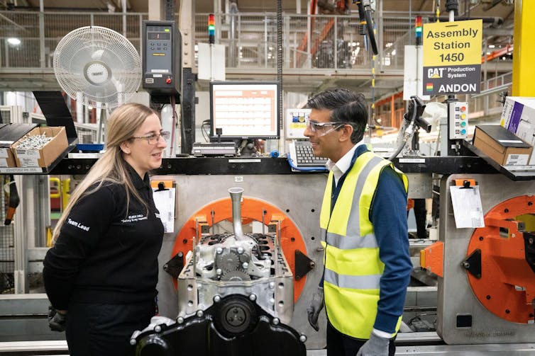 Rishi Sunak wearing a high viz jacket talking to a woman over an engine in a factory.