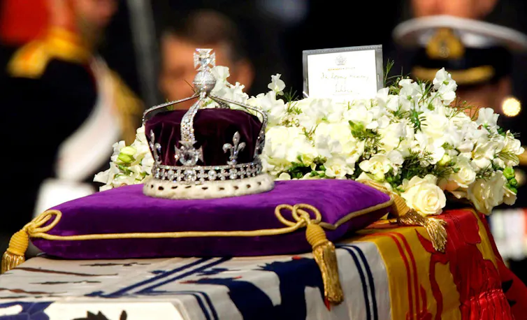 A crown with the large Koh-i-noor diamond sits on a purple cushion, on top of a coffin draped in flags
