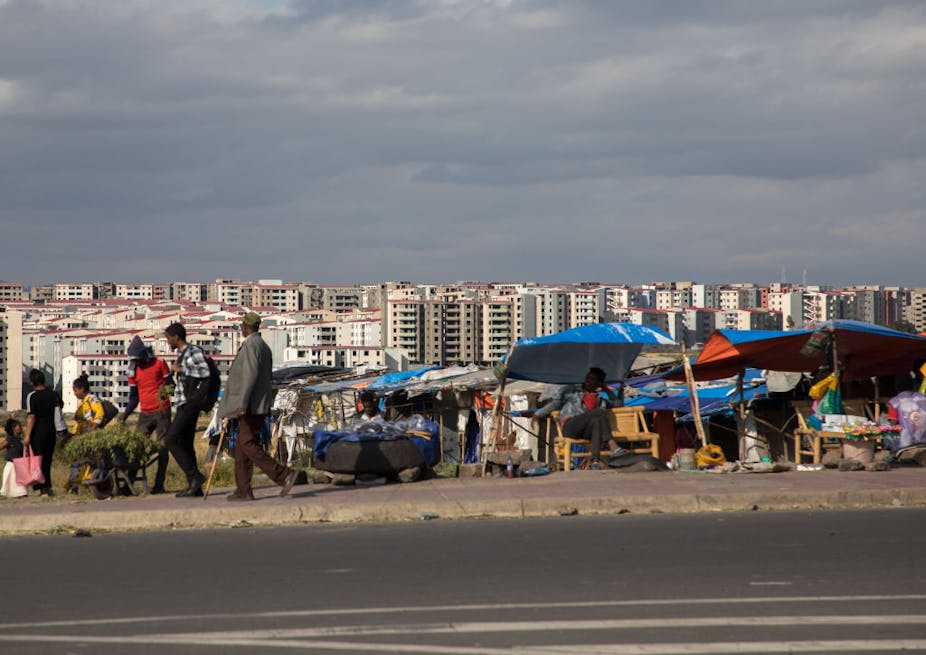 Several high-rise apartments are seen in the background as people walk past street sellers on a busy pavement.