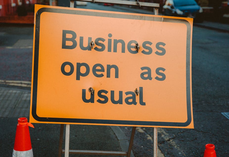 Orange construction sign stating "business open as usual", on a road with cars in the background and two traffic cones in the foreground.