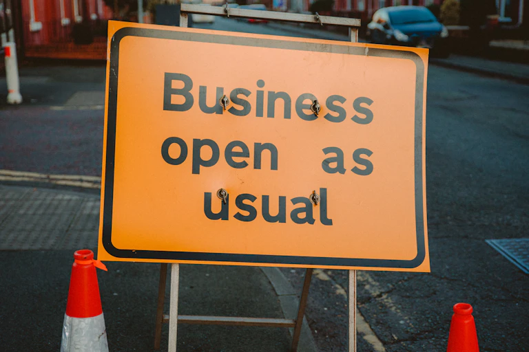 Orange construction sign stating "business open as usual", on a road with cars in the background and two traffic cones in the foreground.