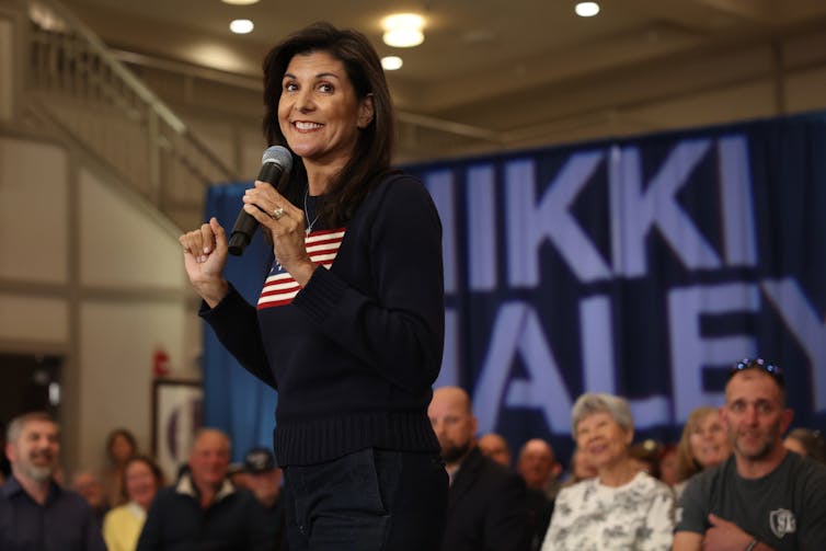 A woman stands on a stage holding a microphone surrounded by people sitting on chairs.