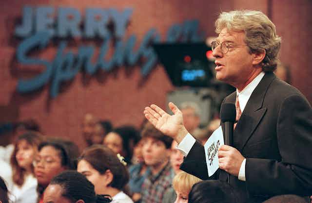 Man wearing glasses holds microphone while gesturing.