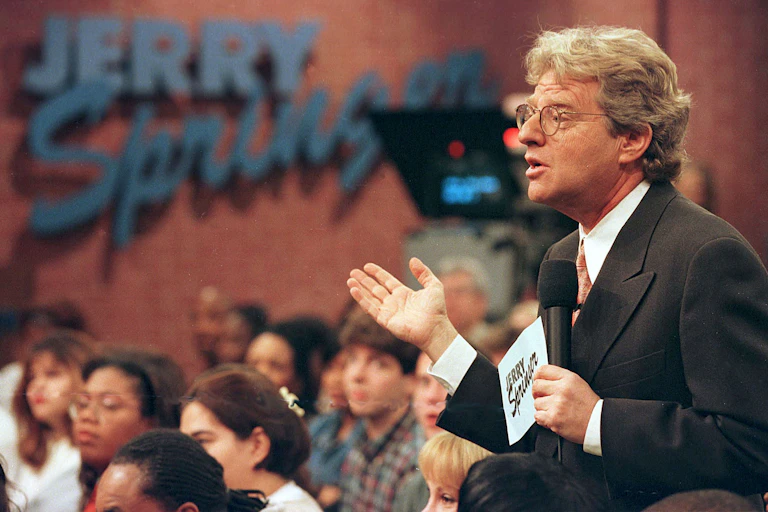 Man wearing glasses holds microphone while gesturing.