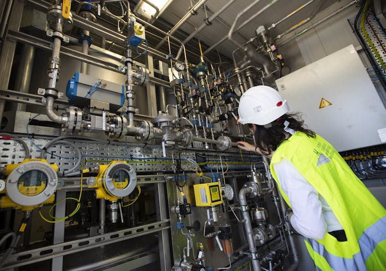 A woman in a work vest adjusts a mechanical system with pipes on a small wall.