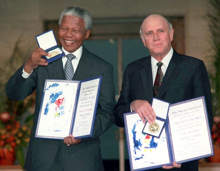 a Black man and a white man stand next to each other holding Nobel Peace prize folders and medals