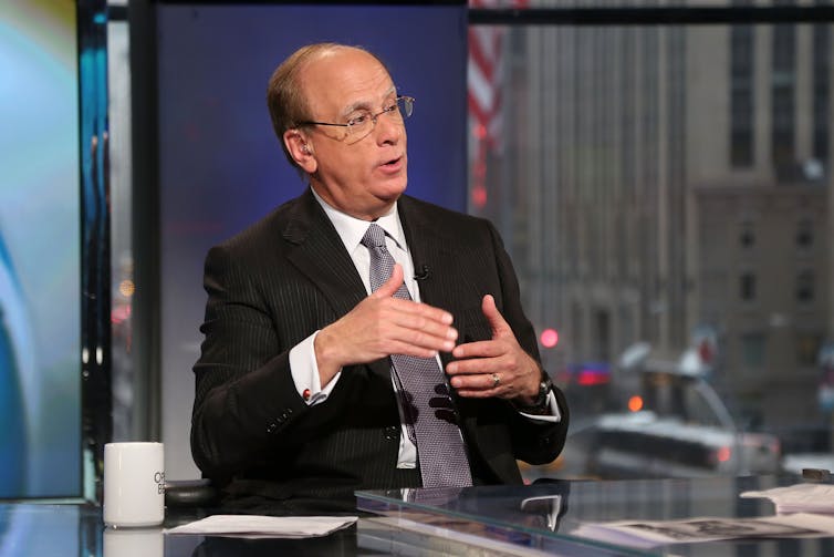 Lark Fink, in business attire and glasses, sits in a news studio being interviewed.