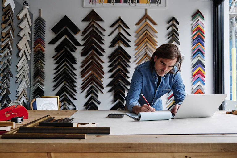 A white man wearing a blue jean shirt uses a pencil to write in a notebook in front of a laptop on a desk with an assortment of frame models on the wall behind him
