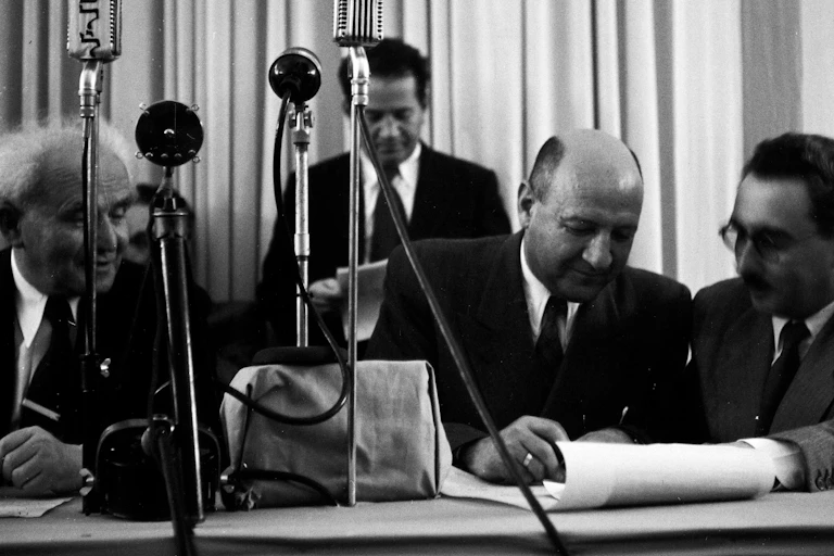 Israel declares independence. Left to right David Ben Gurion, Moshe Kaplan and Moshe Sharett sign the declaration in front of microphones.