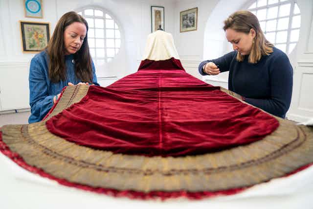 Two women work on a red velvet cape.